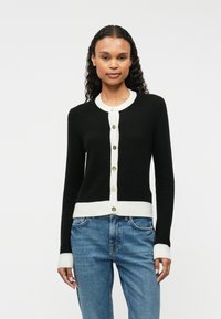 Cardigan noir avec un col et un ourlet côtelés blancs, comportant six boutons verts sur le devant. Texture douce et design ajusté.