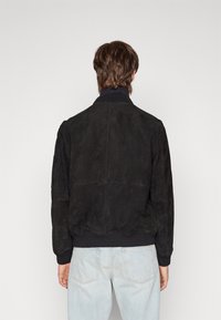 Giubbotto bomber in suede nero con colletto e polsini a coste. Presenta una superficie liscia e un design semplice, con cuciture minime e senza loghi visibili.