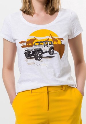 T-shirt blanc avec un motif représentant un jeep blanc, un coucher de soleil orange, des éléphants, des girafes et des acacias, porté avec un pantalon jaune.