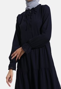 Mørkeblå kjole med lagdelt design, elastiske manchetter og snøre ved halsudskæringen. Fremstillet af letvægtsstof.