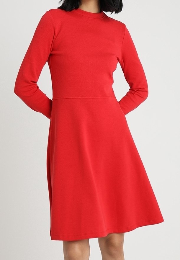 Femme portant une robe rouge longueur genoux à manches longues, avec un corsage ajusté et une jupe évasée, debout les mains dans le dos.