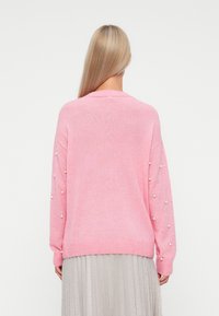 Rosa Strickpullover mit Perlenverzierungen an den Ärmeln, locker geschnitten und mit geripptem Saum. Der Hintergrund ist einfarbig.