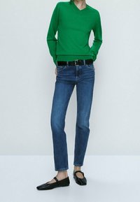 Viher v-kauluksellinen neulepaita, joka on yhdistetty sinisiin farkkuihin ja mustiin tasapohjaisiin kengiin. Neulepaita on istuva ja farkut ovat suoralahkeiset.