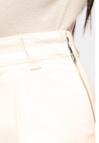 Κοντινό πλάνο σε παντελόνι εκρού με πλευρικό φερμουάρ και θηλιά για ζώνη, φορεμένο με μπεζ πλεκτό τοπ.