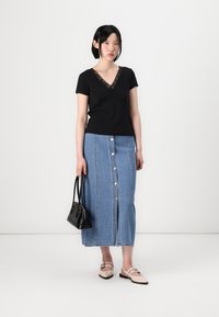 Haut noir à manches courtes avec détail en dentelle au niveau du col, associé à une jupe en denim bleu clair avec des boutons sur le devant et une fente. Sac à main noir et ballerines beiges.