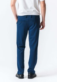 Blå bukser med en glat, elegant tekstur, der har to baglommer med knapper og en lige ben-design. Båret med sorte sko.