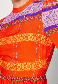 Robe orange vif avec un tissu à motifs en rayures violettes, jaunes et vertes. Avec des manches bouffantes et une taille froncée, elle présente des détails complexes.