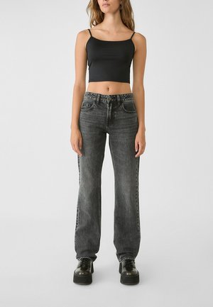 Vrouw met een zwart cropped tanktop, hooggetailleerde vervaagde zwarte spijkerbroek en zwarte plateauzolen, staand tegen een effen grijze achtergrond.