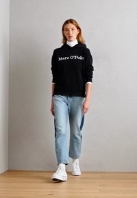 Schwarzer Hoodie mit "Marc O'Polo"-Druck, kombiniert mit hellblauen Jeans und weißen Sneakers, stehend auf einem Holzboden vor einer neutralen Wand.