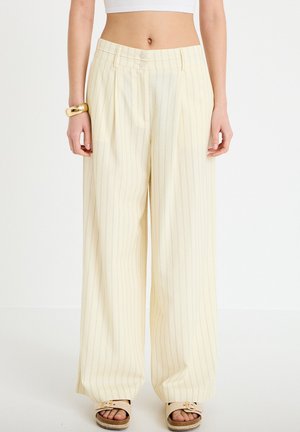 Kvinde iført cremefarvede bredbenede bukser med højtaljet pasform og tynde lodrette striber, kombineret med åbne sandaler og et gyldent armbånd på venstre håndled.