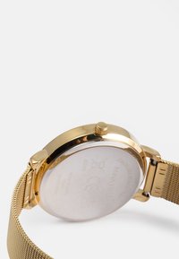 Orologio con cinturino in maglia dorata, caratterizzato da una cassa rotonda, retro in acciaio inossidabile e una corona prominente. Superficie liscia con una finitura riflettente.