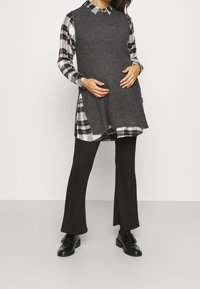 Grijze gebreide vest over een geruite overhemden, gecombineerd met zwarte uitlopende broeken en zwarte schoenen. Het vest heeft zijsplitten voor extra bewegingsvrijheid.