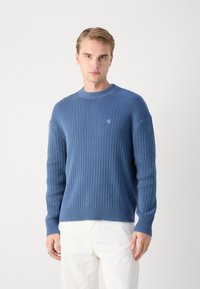Sweter z prążkowanej dzianiny w kolorze niebieskim, z okrągłym dekoltem i długimi rękawami. Małe logo na piersiach. Noszony z białymi spodniami.