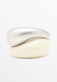 Anelli impilati; il superiore è di un metallo argentato con una superficie liscia, mentre quello inferiore è di un avorio opaco con una forma arrotondata. Entrambi presentano un design minimalista.