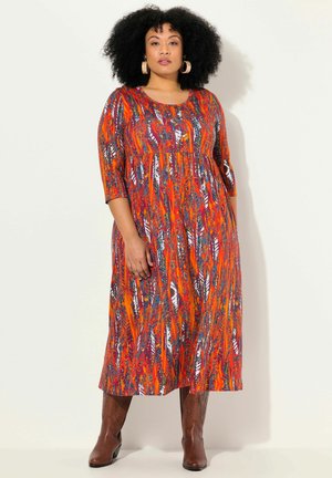 Maxikleid mit rot-blauem Muster, 3/4-Ärmeln, runder Halslinie und tailliertem Oberteil. Tragen mit braunen kniehohen Stiefeln.