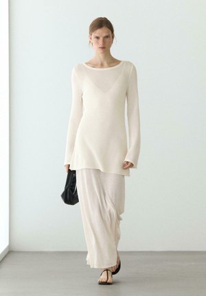 Donna che cammina al chiuso indossando una maglia bianca trasparente a maniche lunghe, una gonna bianca fluente, sandali neri e tenendo una piccola borsa nera.