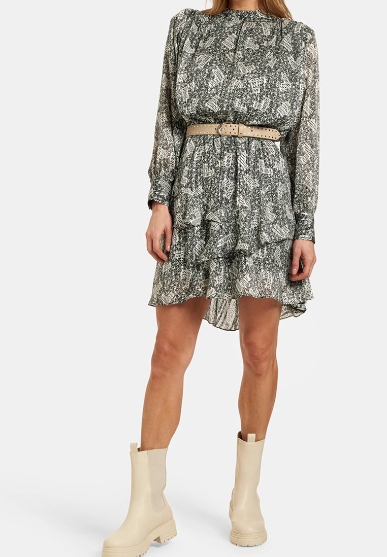 Femme portant une robe à manches longues à motif noir et blanc avec une ceinture beige et des bottines crème, debout sur un fond uni.