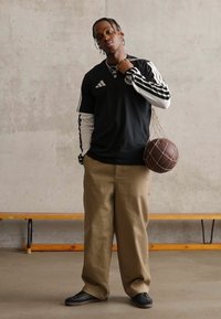 Νεαρός άνδρας με μαύρα αθλητικά ρούχα Adidas και μπεζ παντελόνι, κρατάει καφέ μπάλα μπάσκετ σε δίχτυ τσάντας στον ώμο του, μπροστά σε απλό τοίχο.