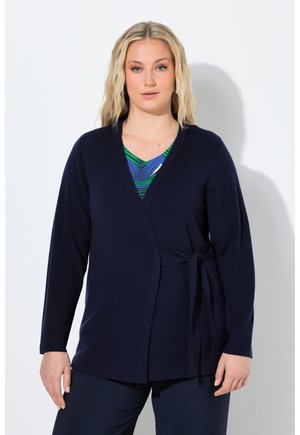 Blonde Frau, die eine marineblaue Wickelstrickjacke über einem grün-blau gemusterten Oberteil trägt und vor einem weißen Hintergrund steht.