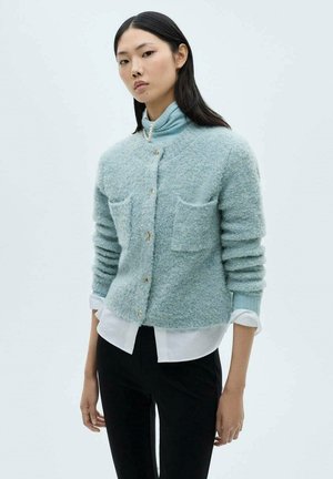 Light blue, textured cardigan with two front pockets, gold buttons, and a cropped silhouette. Worn over a white shirt and black pants.