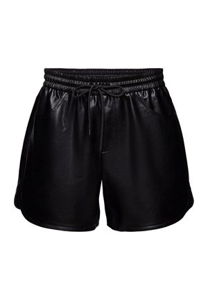 Femme portant un blazer à carreaux, une chemise blanche, un short en cuir noir, des bottes noires montantes et tenant un petit sac à main rouge foncé.