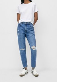 Jeans descoloridos de tiro alto con detalles desgastados y un ajuste relajado, combinados con una camiseta blanca simple y zapatillas de corte bajo.