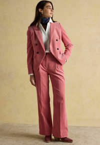 Rozā kordurojas kostīms ar dubultpogājamu žaketi un platajiem bikšu kājiņām, kopā ar baltu kreklu un rakstainu šalli. Brūnas augstpapēžu zābaki.