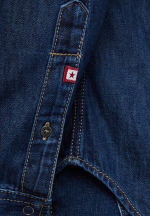 Tela vaquera con costuras dobles amarillas visibles, una pequeña etiqueta roja y blanca con una estrella roja, y un botón a presión negro en una prenda.