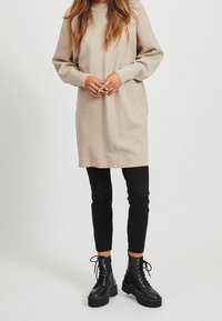Abito in maglia beige con collo alto, maniche lunghe e vestibilità morbida, abbinato a pantaloni neri aderenti e stivaletti neri con lacci spessi.