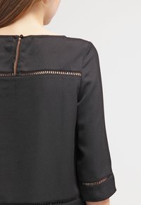 Blusa nera con apertura rettangolare sulla schiena dotata di bottone, pannelli trasparenti sulle spalle e tessuto strutturato.