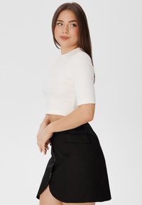 Bílý žebrovaný crop top s krátkými rukávy, kombinovaný s černou sukní na knoflíky se bočním rozparkem. Jednoduchý design a čisté barvy.