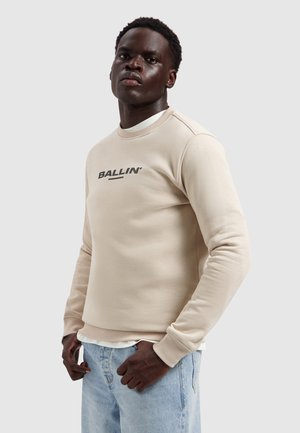 Man in beige sweatshirt met de tekst "BALLIN'" en lichtblauwe jeans, staand tegen een effen lichtgrijze achtergrond.
