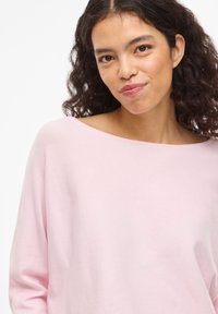 Jauna sieviete ar cirtainiem tumšiem matiem, valkājot gaiši rozā krāsas džemperi bez plecu daļas, maigi smaida pret vienkrāsainu baltu fonu.