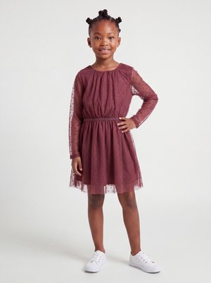 Jeune fille aux cheveux tressés portant une robe bordeaux à manches longues avec un voile à pois et des baskets blanches, souriant avec une main sur la hanche.