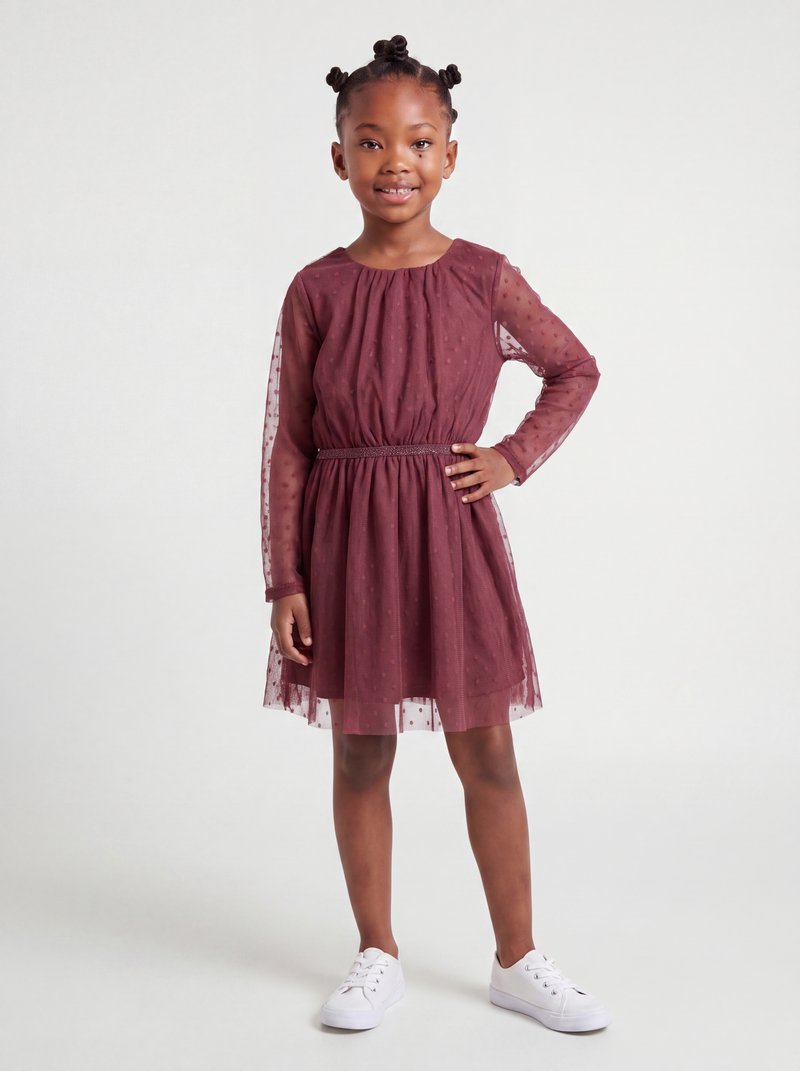 Jeune fille aux cheveux tressés portant une robe bordeaux à manches longues avec un voile à pois et des baskets blanches, souriant avec une main sur la hanche.