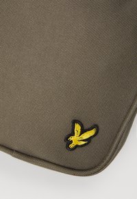 Geantă din țesătură de culoare verde măsliniu, cu o suprafață texturată și un logo cu un păun galben cu margini negre. Are o formă rotundă și un detaliu vizibil de cusătură.