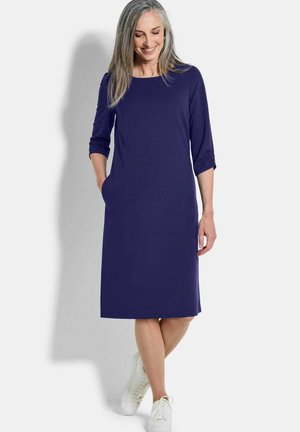 Frau mit langen grauen Haaren, die ein knielanges dunkelblaues Kleid mit 3/4-Ärmeln und weißen Turnschuhen trägt, steht vor einem hellen Hintergrund.