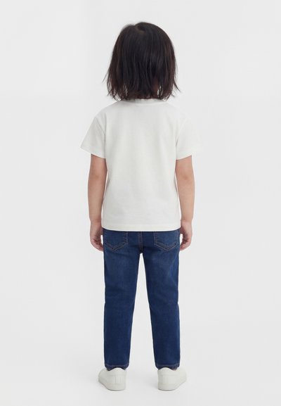 Vállig érő sötét hajú gyermek, háttal állva, egyszerű fehér rövid ujjú pólót, kék farmert és fehér sportcipőt visel.