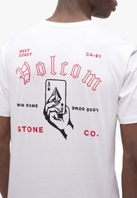 Homme portant un t-shirt blanc avec un graphisme rouge et noir d'une main tenant l'as de pique et le texte "Volcom Stone Co." dans le dos.