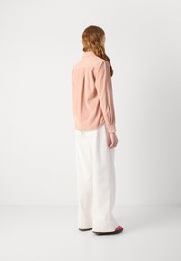 Light pink long-sleeve shirt with a collar, featuring a back pleat. Paired with white wide-leg trousers and pink-accented shoes.