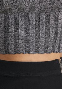 Detalle de un top corto de punto acanalado en gris y negro, con el dobladillo por encima de unos pantalones negros que tienen una cremallera metálica visible en el lado.