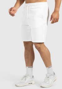 Weiße Sportshorts aus Baumwollmischung mit einem elastischen Bund, seitlichen Taschen und einer lockeren Passform, kombiniert mit weißen Sneakers und Sportsocken.