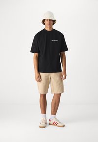 Camiseta negra de algodón con texto blanco, pantalones cortos beige, sombrero tipo cubo blanco, zapatos rojos y beige. El modelo está de pie sobre un fondo liso.