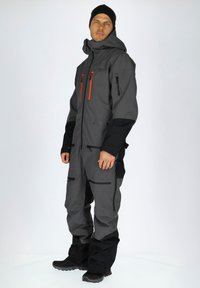 Grijs geïsoleerd overall met zwarte accenten, voorzien van ritszakken, een hoge kraag en een verstelbare capuchon. Duurzame stof met een matte afwerking.