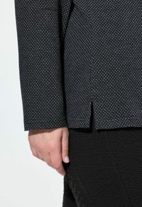 Mano rilassata accanto al corpo, che indossa pantaloni neri con texture e un top a maniche lunghe scuro con un piccolo spacco laterale.