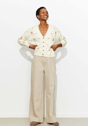 Personne souriante portant un cardigan crème à fleurs boutonné, un pantalon large beige et des mocassins marron, debout les mains sur les hanches.