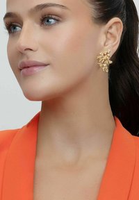 Guldörhängen har en abstrakt, grenliknande design med texturerade accenter. Modellen bär en orange kavaj som tydligt visar örhängena.