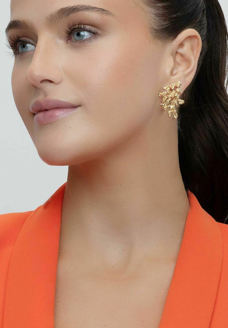 Guldörhängen har en abstrakt, grenliknande design med texturerade accenter. Modellen bär en orange kavaj som tydligt visar örhängena.