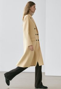 Langer, doppelreihiger Mantel aus weichem gelbem Stoff mit schwarzen Knöpfen, kombiniert mit eng anliegenden dunkelbraunen Hosen und schwarzem Schuhwerk.