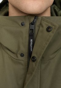Olive green jacket with a zip and snap button closure. Features a textured fabric and a logo tag reading "REVOLUTION." Close-up view of neck area.